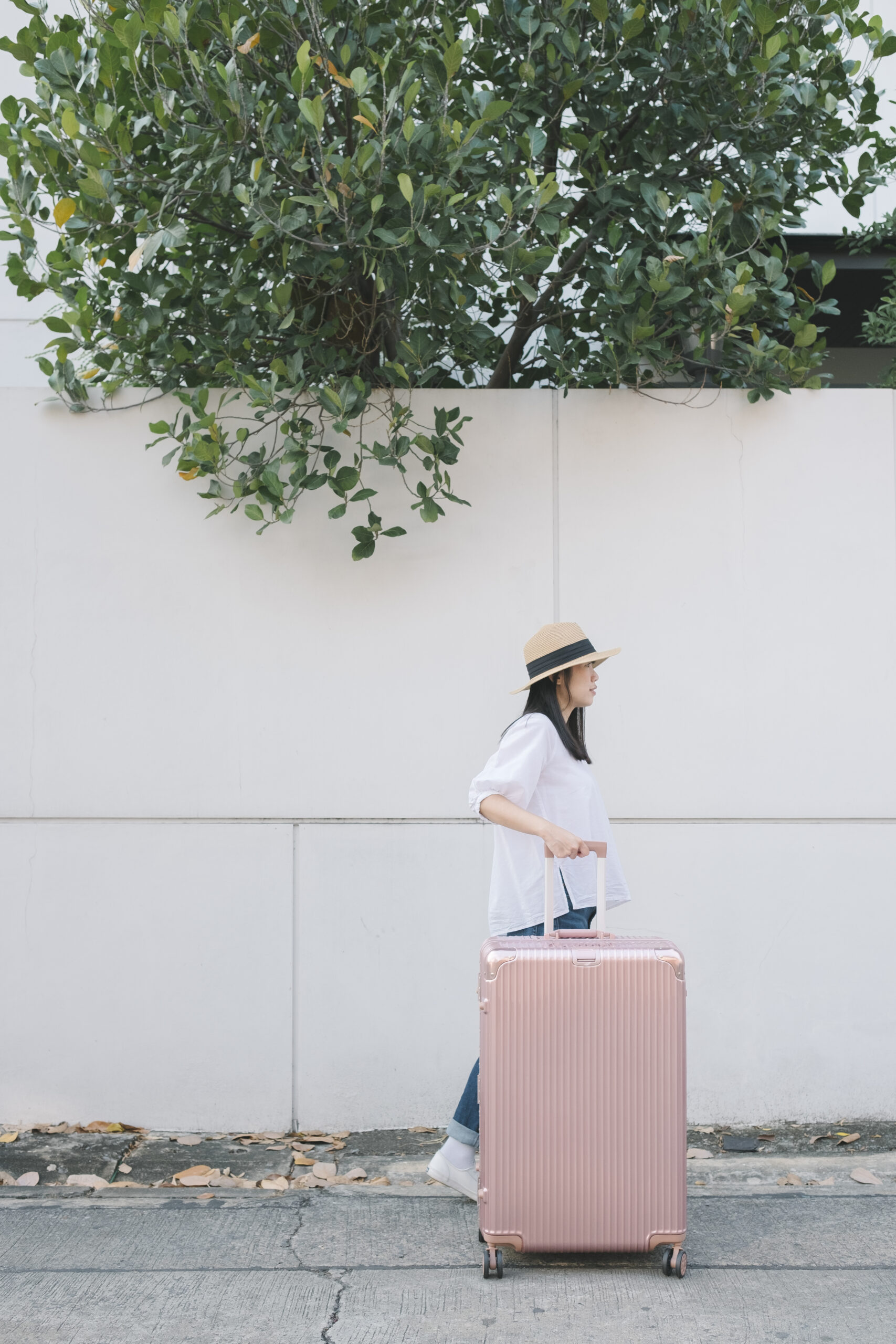 Woman wearing a hat and white shirt walking with a large pink suitcase beside a white wall with green leafy tree above.